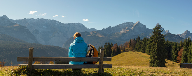 A women/ hiker is seen from her back, sitting on a bench in front of a sunny alpine mountain panorama in fall, close to Garmisch-Partenkirchen. The peak visible to the right is the Zugspitze, Germany's highest mountain peak.