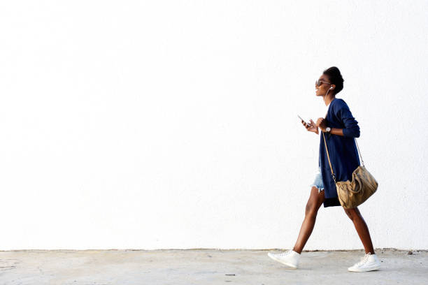 Trendy black woman listening music on mobile phone Full length side view portrait of trendy young black woman walking outdoors and listening to music on her mobile phone walking stock pictures, royalty-free photos & images