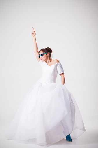 Full length portrait of nerdy bride wearing sunglasses. She is dancing and holding her right arm raised