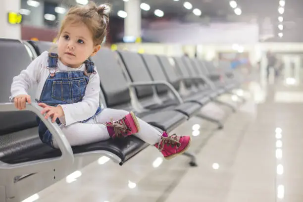 Pretty 20 months old baby girl sitting alone on an airport seat in waiting hall, and waiting for the flight to travel with her family .