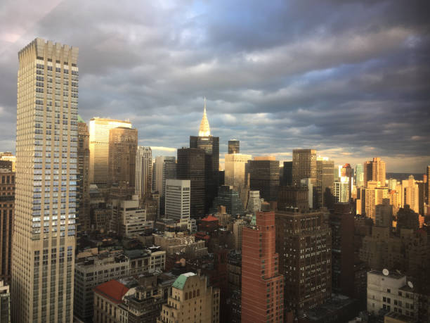 Clouds part and sun shines down on Midtown Manhattan, skyscrapers glowing in the sunlight.  Storm clouds over New York City skyline, taken from tall skyscraper office window. Looking out window.