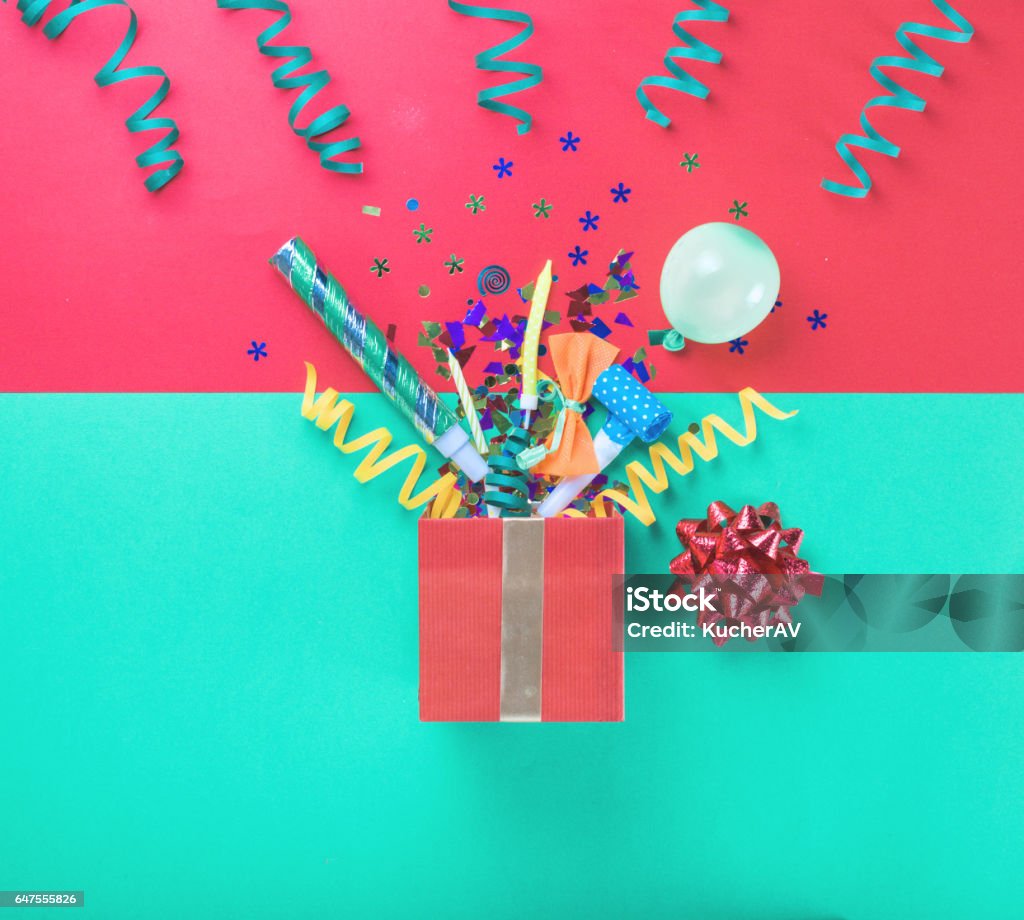 Red gift box with various party confetti, balloons, streamers, noisemakers and decoration on a multicolored background Red gift box with various party confetti, balloons, streamers, noisemakers and decoration on a multicolored background. Top view Gift Box Stock Photo