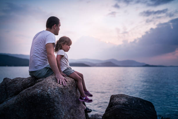 istock Father and daughter sitting on rocks and watching the sunset 647583802