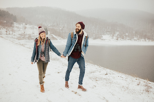 Boyfriend and girlfriend with bright smile on their faces walking on the snowy road near the lake