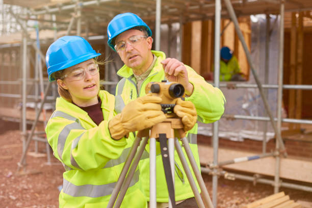 construction worker learning how to use a builder's level a female construction worker stands behind a builder's level on a  building site .Behind her a co-worker walks across the development . civil engineering stock pictures, royalty-free photos & images