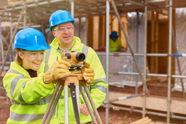 happy female construction trainee with tutor a female construction worker stands behind a builder's level on a  building site .Behind her a co-worker walks across the development . theodolite photos stock pictures, royalty-free photos & images