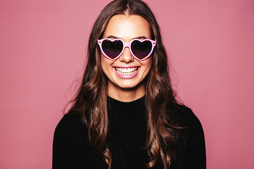 Portrait of beautiful young woman with heart shaped sunglasses and smiling against pink background. Caucasian fashion model posing with glasses.