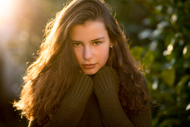 Teenage girl looking shy at camera Outdoors portrait of a teenage girl with curly long brown hair in front of some leaves. She is looking serious at the camera wearing a green pullover. jugendkultur stock pictures, royalty-free photos & images