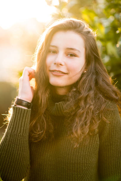 Teenage girl smiling happy at camera Outdoors portrait of a teenage girl with curly long brown hair in front of some leaves. She is looking friendly at the camera wearing a green pullover. There are some lens flares from the back light. jugendkultur stock pictures, royalty-free photos & images