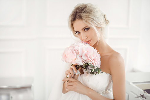 Classical young gourgeous bride. Studio interior fashion shot of fashion model in wedding dress with bouquet of flowers sitting in white room. Blonde woman portait with profeshional make-up and hairstyle.