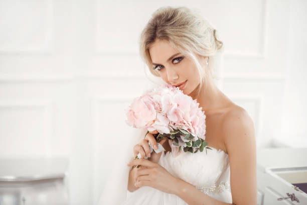 Gourgeous bride studio interior photo Classical young gourgeous bride. Studio interior fashion shot of fashion model in wedding dress with bouquet of flowers sitting in white room. Blonde woman portait with profeshional make-up and hairstyle. wedding dress photos stock pictures, royalty-free photos & images