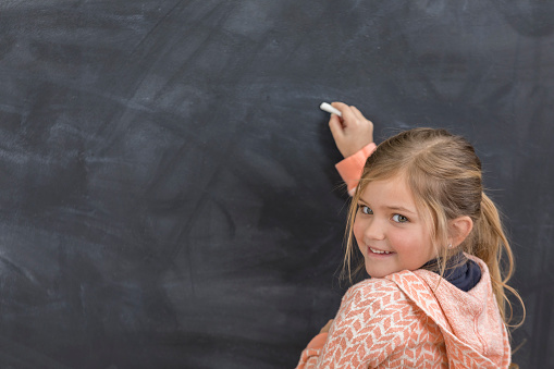 Adorable female kindergarten student writes something on a blackboard at school. She is looking over her shoulder.