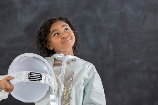 African American elementary schoolgirl dresses as an astronaut for career day at school. She is looking up and standing in front of a chalkboard.