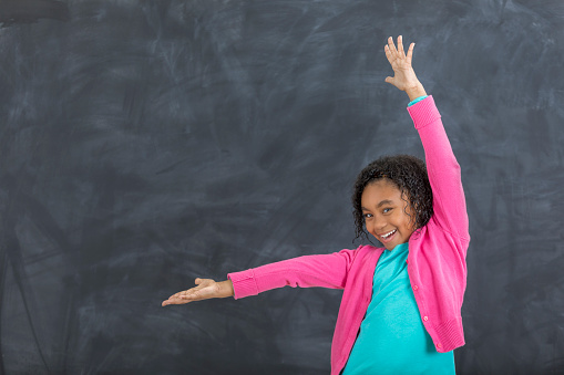 Pretty African American elementary student gestures with her arms in front of a chalkboard in a classroom. She is smiling at the camera.