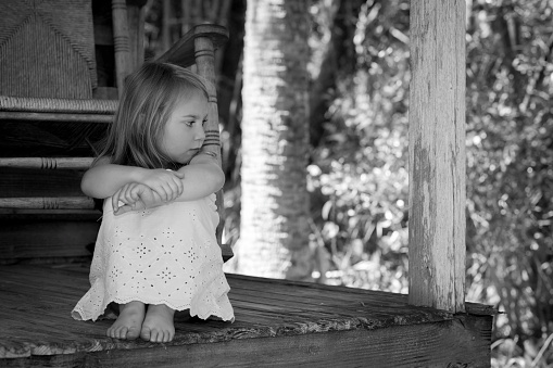 Black and white portrait of a young girl sitting on a porch with her knees tucked up, while looking to the side in a sad manner.