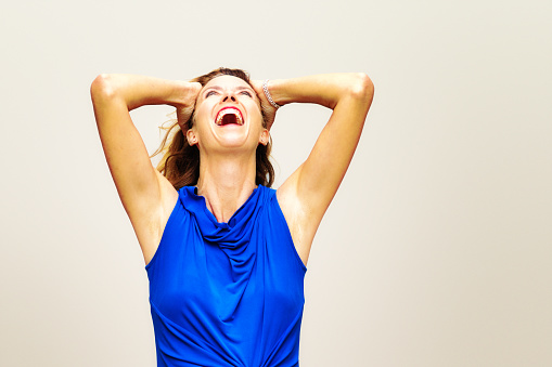 Happy Mature blonde female in blue sleeveless top laughing looking up arms up holding her head.