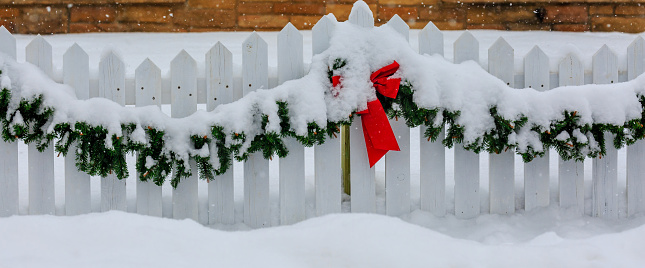 White picket fence decorated for Christmas with red bow and green garland covered with snow.