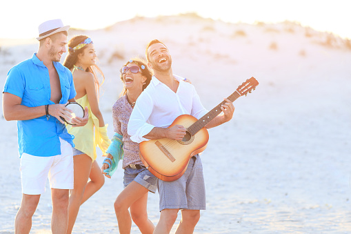Group of young people having fun on beach, dancing and playing guitar and drum. All with casual clothes.