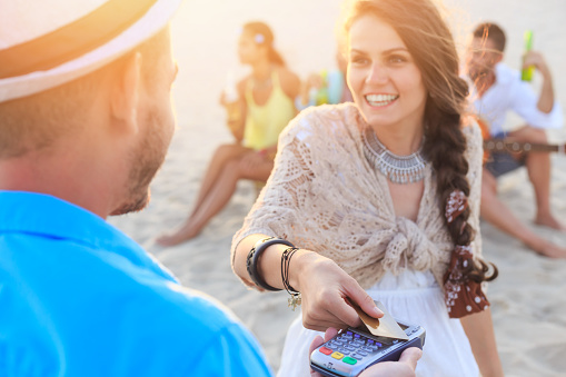 Young woman having fun on beach, using credit card for contactless payment. Wears casual clothes. Friends sitting behind her and drinking beer, playing guitar.