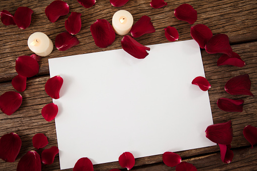 Wax candle and blank paper surrounded with rose petal against wooden background