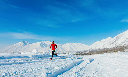A mid adult female wearing winter running wardrobe runs along a rural Mapleton, Utah road training for an upcoming race. The road is completely covered in snow but she is dedicated in her training. Sparse composition.