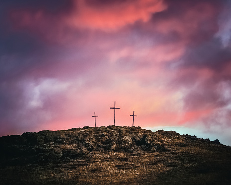Three crosses on top of a rocky hill against a dramatic sky of pink and purple clouds. Symbolic of Easter and Good Friday, holidays celebrated by Christians.