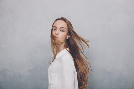 Fashion teenager girl with blowing hair standing against blue textured wall background. Trendy young teen woman in white shirt posing indoors and looking at camera