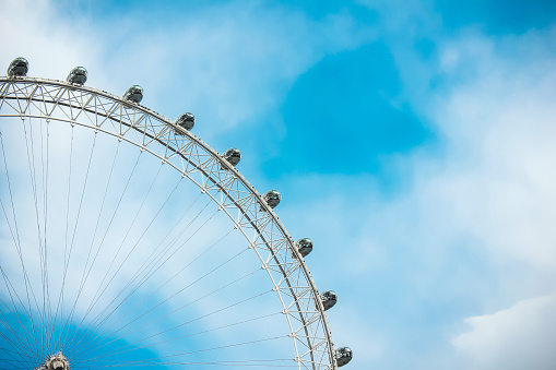 Close up of the London Eye from low angle view. It show a quarter of the ferris wheel, on the left side. The backgrounds is a blue sky with clouds.