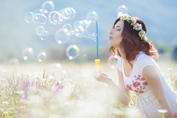 Beautiful pregnant woman with long curly brown hair, dressed in a white T-shirt without sleeves, on his head wearing a wreath of white daisies, spends a summer day in a field of blossoming daisies, blowing bubbles
