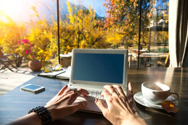 Hands of a young lady freelancer typing on his white laptop on a black wooden table. In the background veranda with glass walls beautiful garden with yellow leaves and mountain. Conceptual Work.