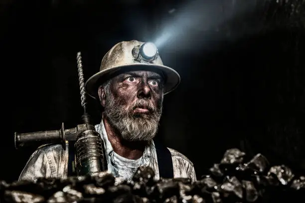 A dirty middle-aged Caucasian male coal miner covered in coal dust with a large hammer drill on his shoulder looking off into the distance, wearing a aluminum hardhat with a LED light attached looking at the camera while in a dark coal mine.