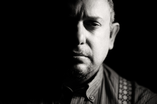 Close-up of a mean-looking man, lit with chiaroscuro, on black background. Only half of his face is illuminated. A mafia boss?