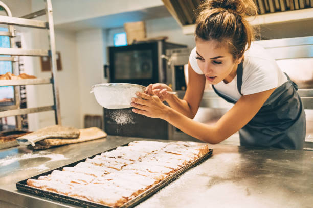 The final touch for the perfect pasrty Smiling young baker dusting pastry with powdered sugar in a commercial kitchen artisanal food and drink stock pictures, royalty-free photos & images