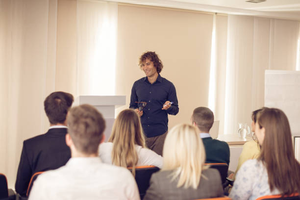 istock Young coach holding speech on conference with many young participants 648736876