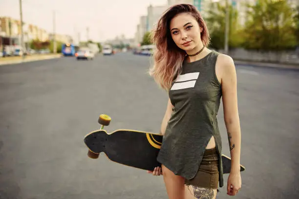Beautiful young tattooed woman with his longboard on the road in the city in sunny weather