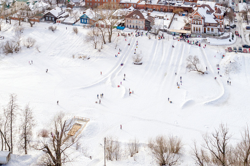 Adults and children are snow tubing and sledding down the hills on the banks of the frozen river in winter frosty day. City Yoshkar-Ola, Mari El Republic, Russia. Bird's-eye view