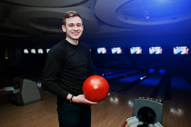 istock Cheerful young man holding a bowling ball and smiling at camera while standing against bowling alleys with ultraviolet light. 648821386