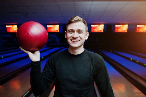 istock Cheerful young man holding a bowling ball and smiling at camera while standing against bowling alleys with ultraviolet light. 648821388