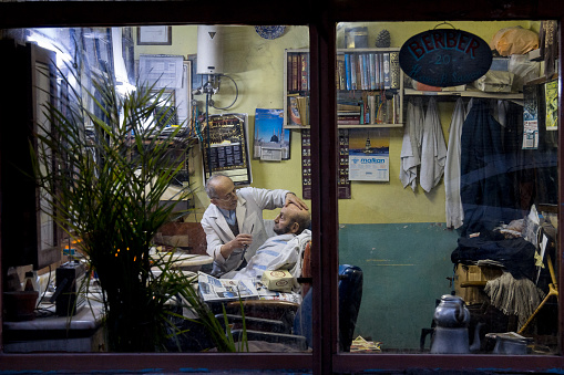 Picture of an old barber and his client taken from outside at dusk