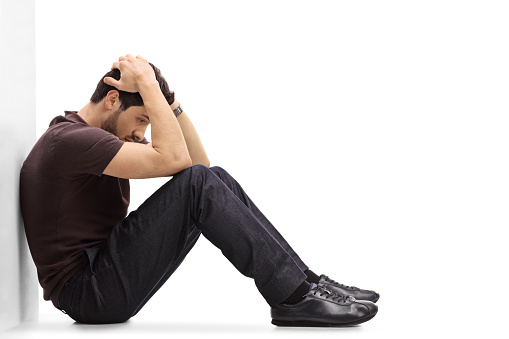 Depressed man sitting on the floor with his head down and leaning against a wall isolated on white background