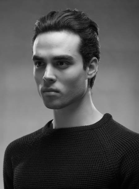 Casual stylish guy posing sitting in the studio on black background. Dressed in a white shirt, blue jeans, a black hat and sneakers