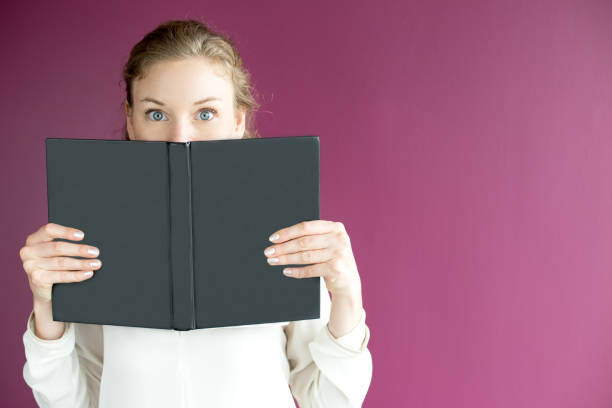 Closeup portrait of young beautiful fair-haired woman looking at camera, holding open book and hiding her face behind it. Isolated front view on purple background.