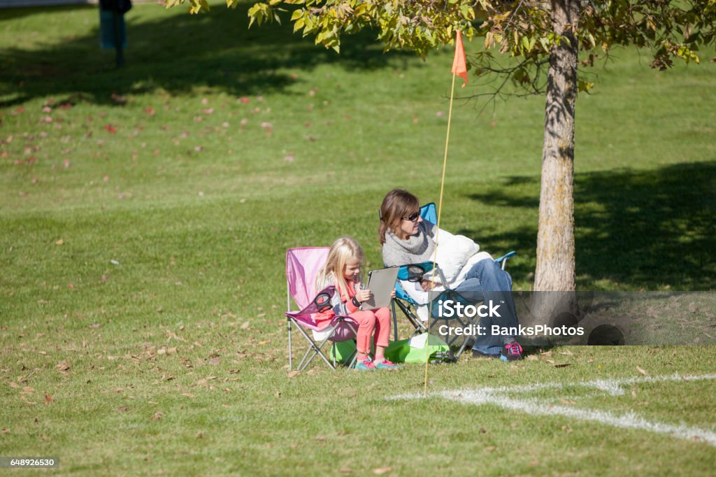 Mother and Child Spectators Sitting on Sideline of Outdoor Field A mother and daughter are sitting along the sideline  watching an outdoor soccer game. The young girl is playing on her electronic device or tablet. Sideline Stock Photo