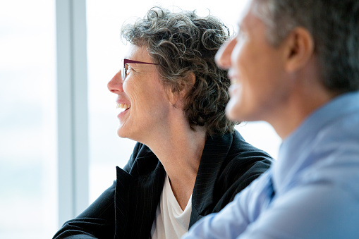 Closeup of smiling businesswoman and businessman attending meeting and looking aside with window in background. Man is blurred. Side view.