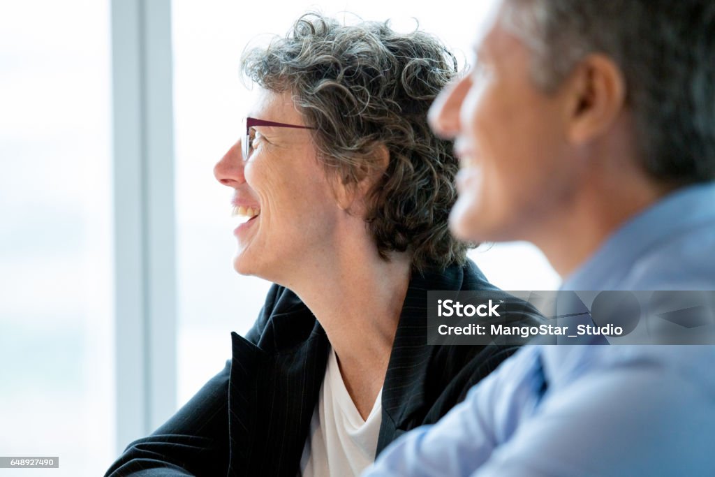 Closeup of Two Smiling Coworkers at Meeting Closeup of smiling businesswoman and businessman attending meeting and looking aside with window in background. Man is blurred. Side view. Candid Stock Photo