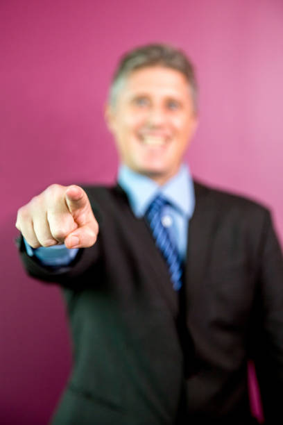 Closeup of smiling middle-aged business man looking straight and pointing at camera with focus on hand. Isolated front view on purple background.
