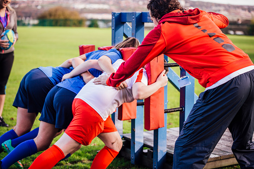 Girls using equipment during rugby training. Using a rack for strength training for scrums they can be seen bent over. Two adults can be seen coaching them.