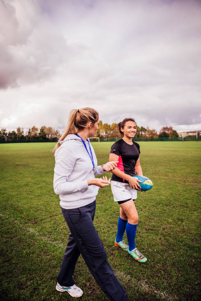 Teenage girl chatting with her coach after rugby training. Holding her rugby ball and covered in mud and dirt as she walks .