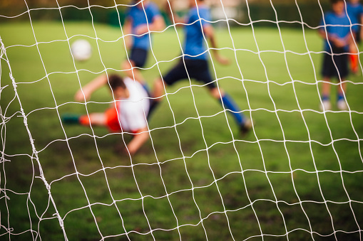 Goalie defending his nets as he dives down while team member kick the ball. Net is in focus and soccer players are blurred