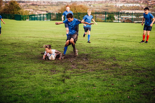Teenage boys playing soccer and one boy falls into the mud in front of the ball as another boy runs in.